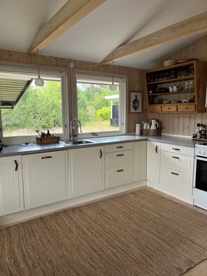A kitchen with white cabinets, a wooden ceiling, a large window, and a built-in shelf with dishes. - Holiday home close to the beach in Nødebohuse (Hundested)