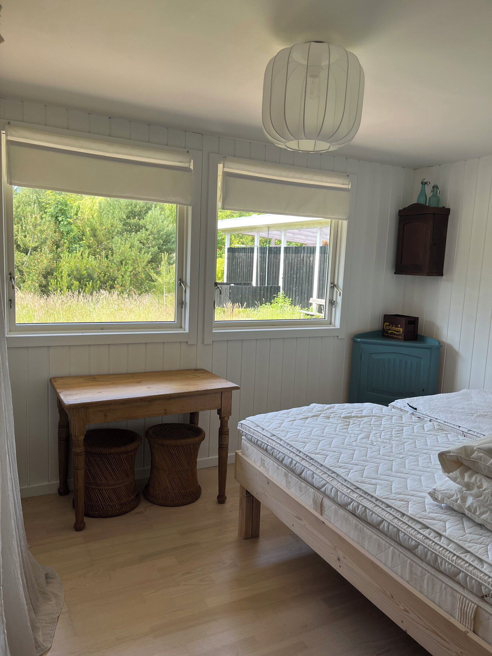 Ein Schlafzimmer mit einem Bett, zwei Fenstern, einem Holztisch, zwei Weidenkörben, einem blauen Schrank und einer Pendelleuchte.
