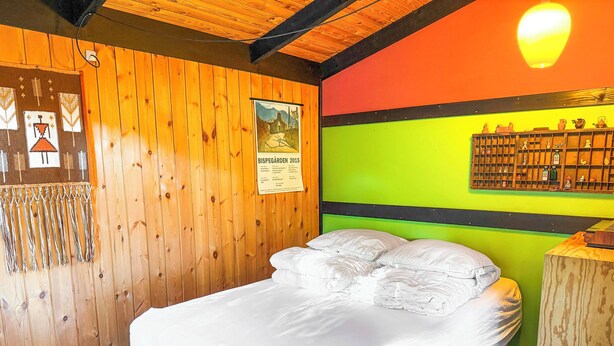 Habitación con pared de madera, cama con ropa de cama blanca, pared verde de acento y estante con diversos objetos.