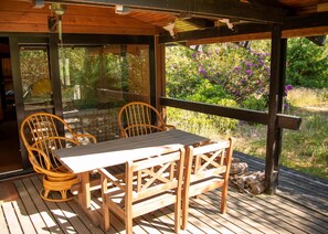 Un área de estar al aire libre con techo, con una mesa y sillas, rodeada de paredes de vidrio y vistas a un jardín con flores moradas.