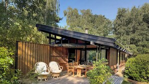 Una terraza de madera con mobiliario exterior y un porche cubierto, con vistas al bosque circundante.