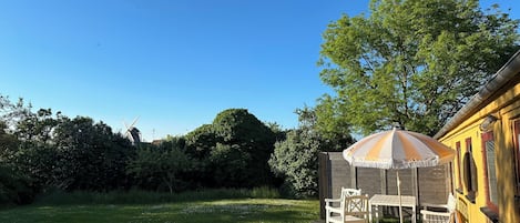 Eine Holzterrasse mit Gartenmöbeln, einem gestreiften Sonnenschirm und einem gelben Gebäude mit Fenstern.