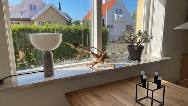 A dining area with a wooden table, a window with a view of houses, a potted plant, and a decorative piece on the windowsill. - Holiday Flat in Skagen town (Skagen)