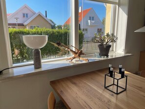 Un comedor con una mesa de madera, un ventanal con vistas a casas, una planta en maceta y un adorno en el alféizar.