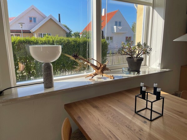 A dining area with a wooden table, a window with a view of houses, a potted plant, and a decorative piece on the windowsill.