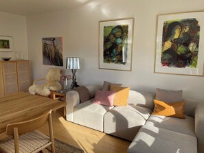Una sala de estar con un sofá, una mesa de madera y una lámpara. Hay cuadros abstractos en la pared.