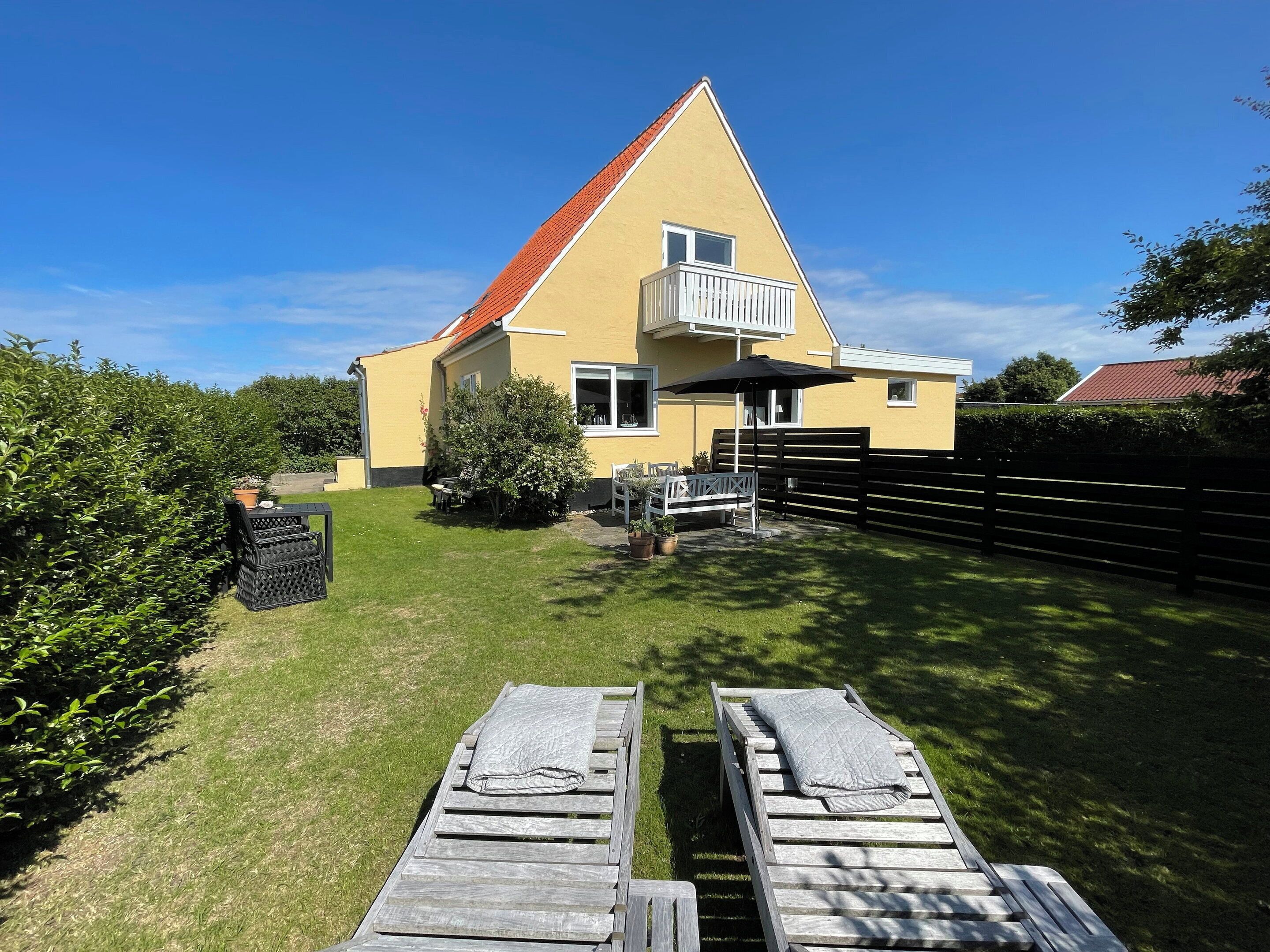 Et gult hus med rødt tag, en balkon og en træterrasse med liggestole.