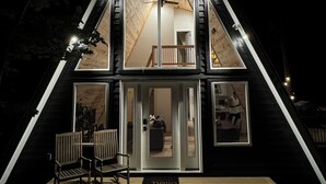 A triangular wooden cabin with a balcony, a door with a 'hello' mat, and two chairs outside.