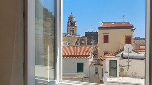 Vista pela janela de um edifício com uma torre sineira, telhados de telha vermelha e antenas parabólicas.