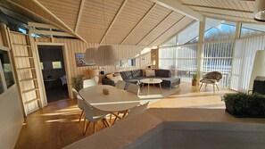 A spacious living area with a high ceiling, wooden beams, a sofa set, a dining table, and a large window providing natural light.
