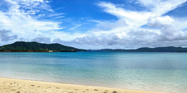 Una playa con agua turquesa cristalina, una orilla arenosa y una cadena montañosa distante bajo un cielo parcialmente nublado.
