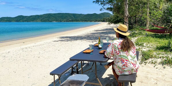 A beachside table set with a bottle, glasses, and a menu, overlooking a sandy beach and turquoise waters. - Le Grand Bleu (Tatsugo)