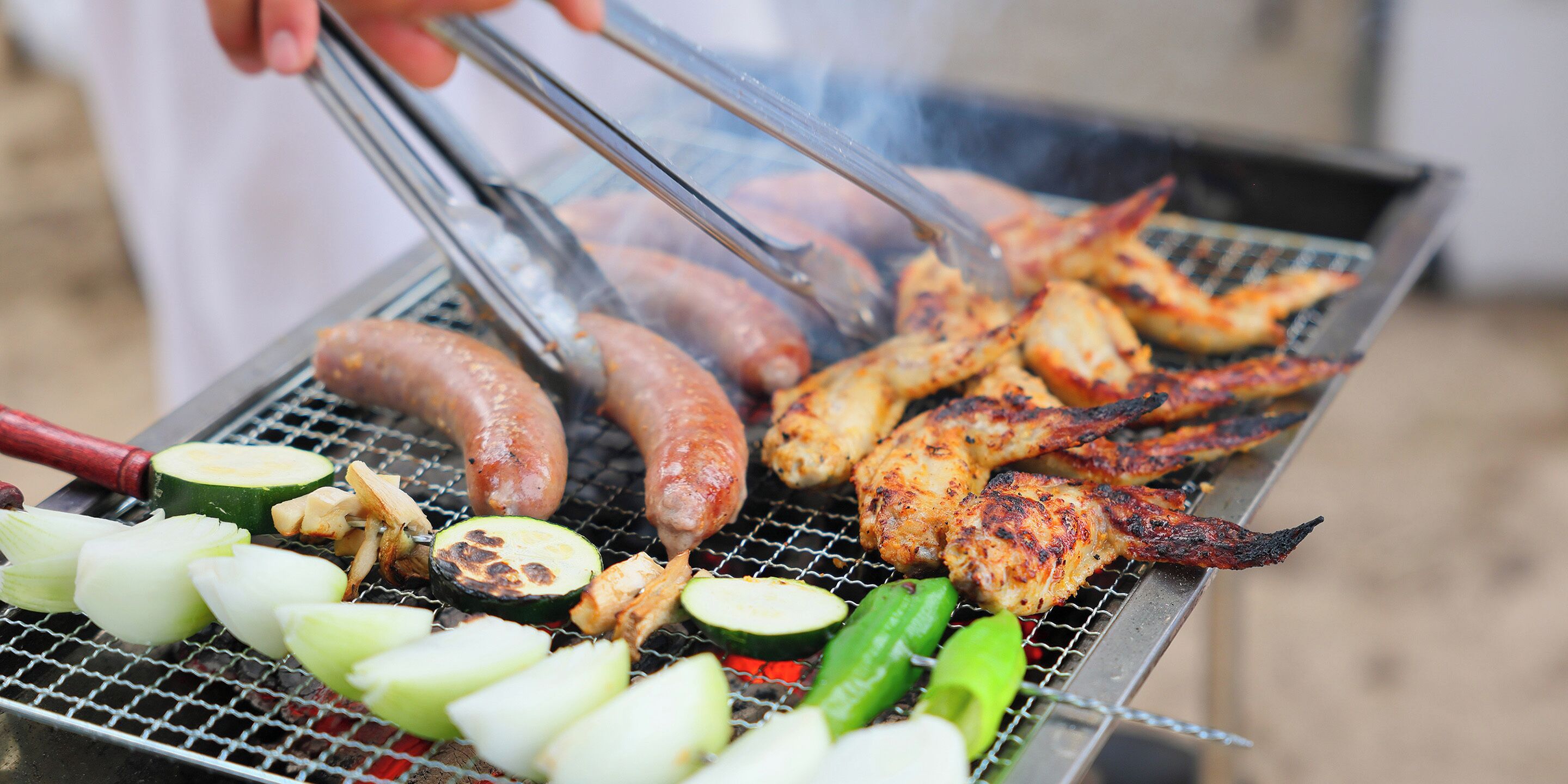 Una parrilla con salchichas, alitas de pollo, calabacín y otras verduras asándose.