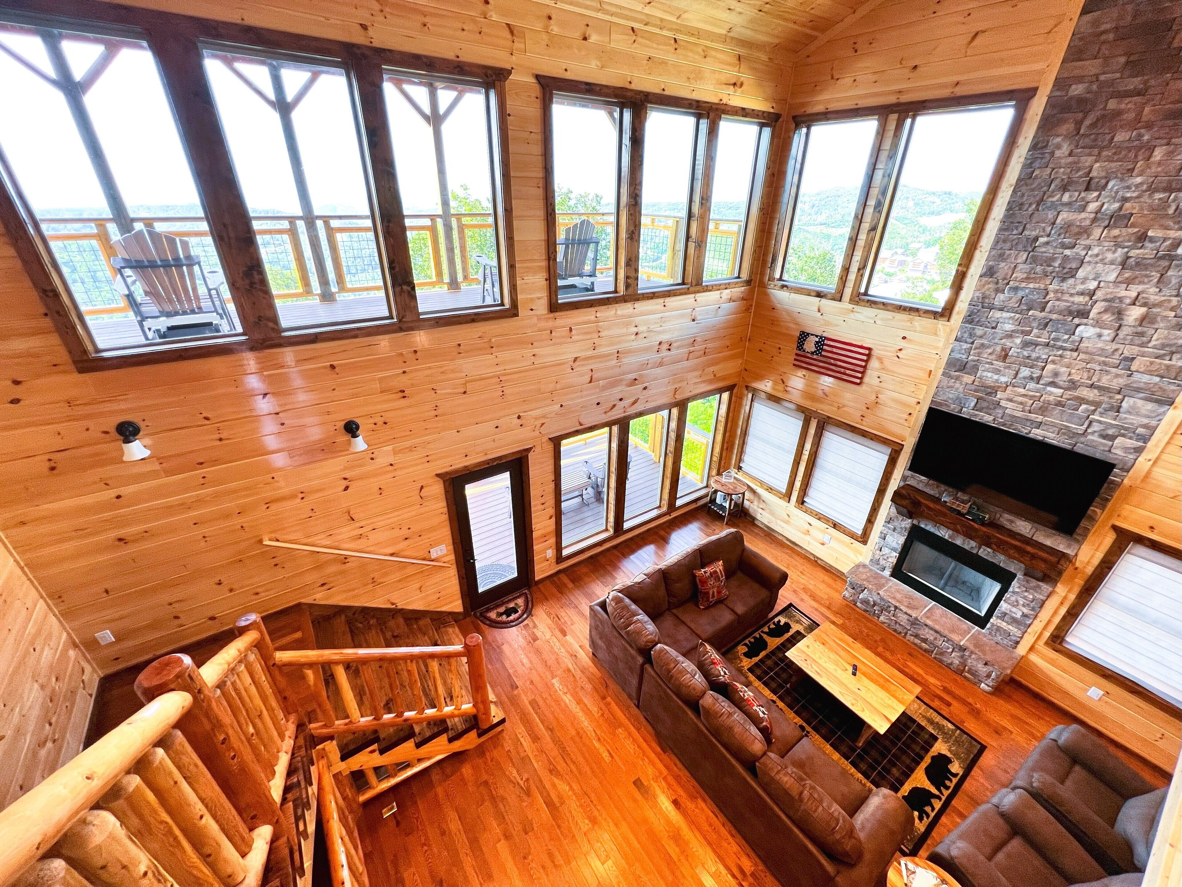 A spacious living area with a large wooden staircase, a stone fireplace, and multiple large windows offering views of the surrounding landscape.