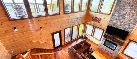 A spacious living area with a large wooden staircase, a stone fireplace, and multiple large windows offering views of the surrounding landscape.