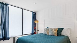 Quarto com cama grande, janela com cortinas, mesa de cabeceira com abajur e radiador fixado na parede.