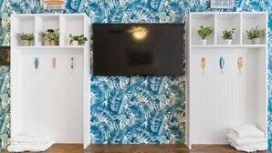 Quarto com uma televisão de tela grande fixada na parede, bancos de madeira e prateleiras com plantas e pranchas de surf.