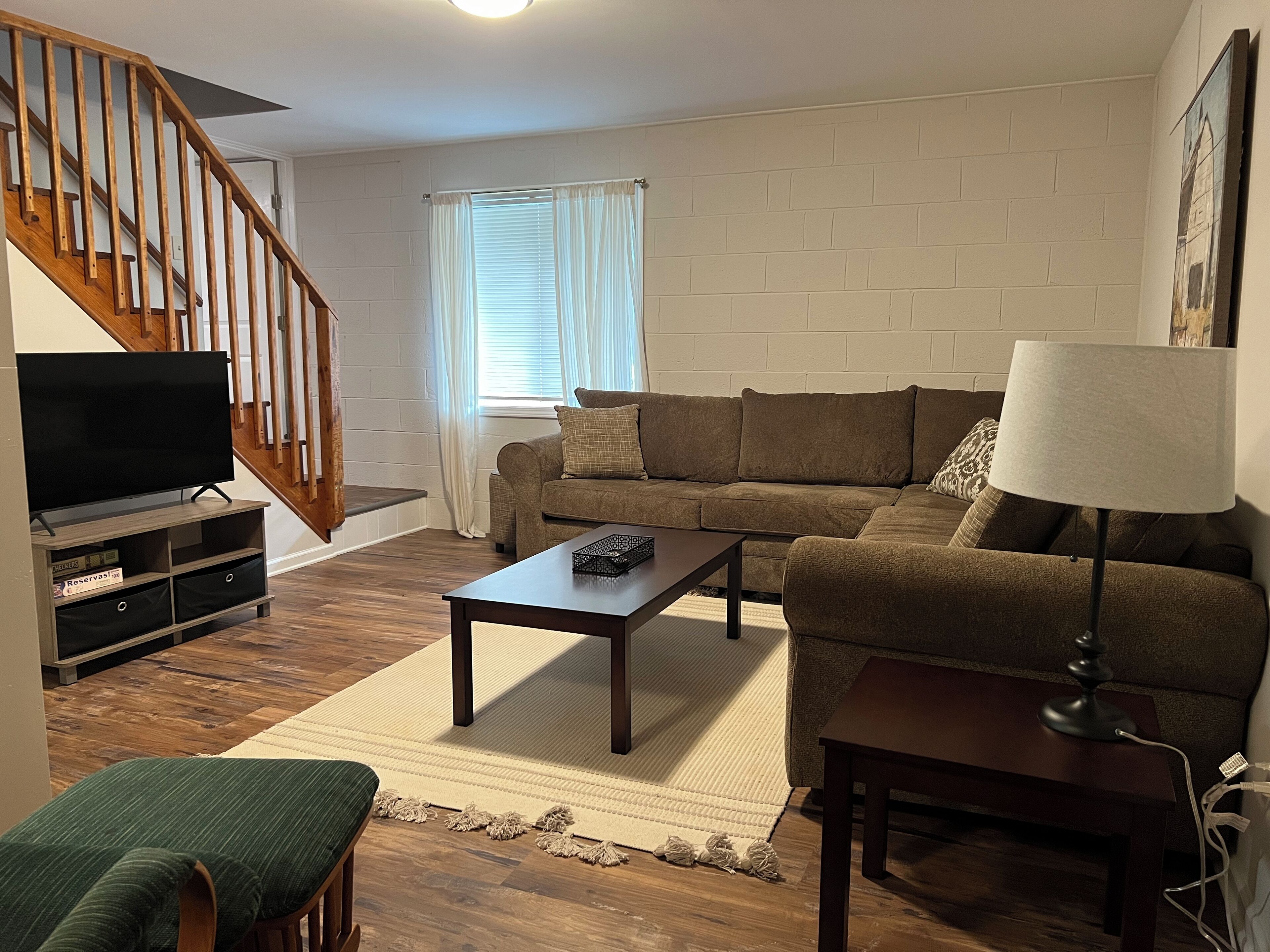 A living room with a sofa, a coffee table, a TV on a stand, a lamp, a staircase, and a window with curtains.