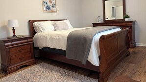 Een slaapkamer met een houten bed, nachtkastje met lamp, een dressoir, vloerkleed, schilderij en spiegel.