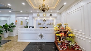 A hotel reception desk with a chandelier, a reception counter, and a decorative display with flowers and fruits. - Senior Hotel (Can Tho)