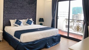 A hotel room with a large bed, a nightstand, a lamp, and a view of the city through a balcony. - Senior Hotel (Can Tho)