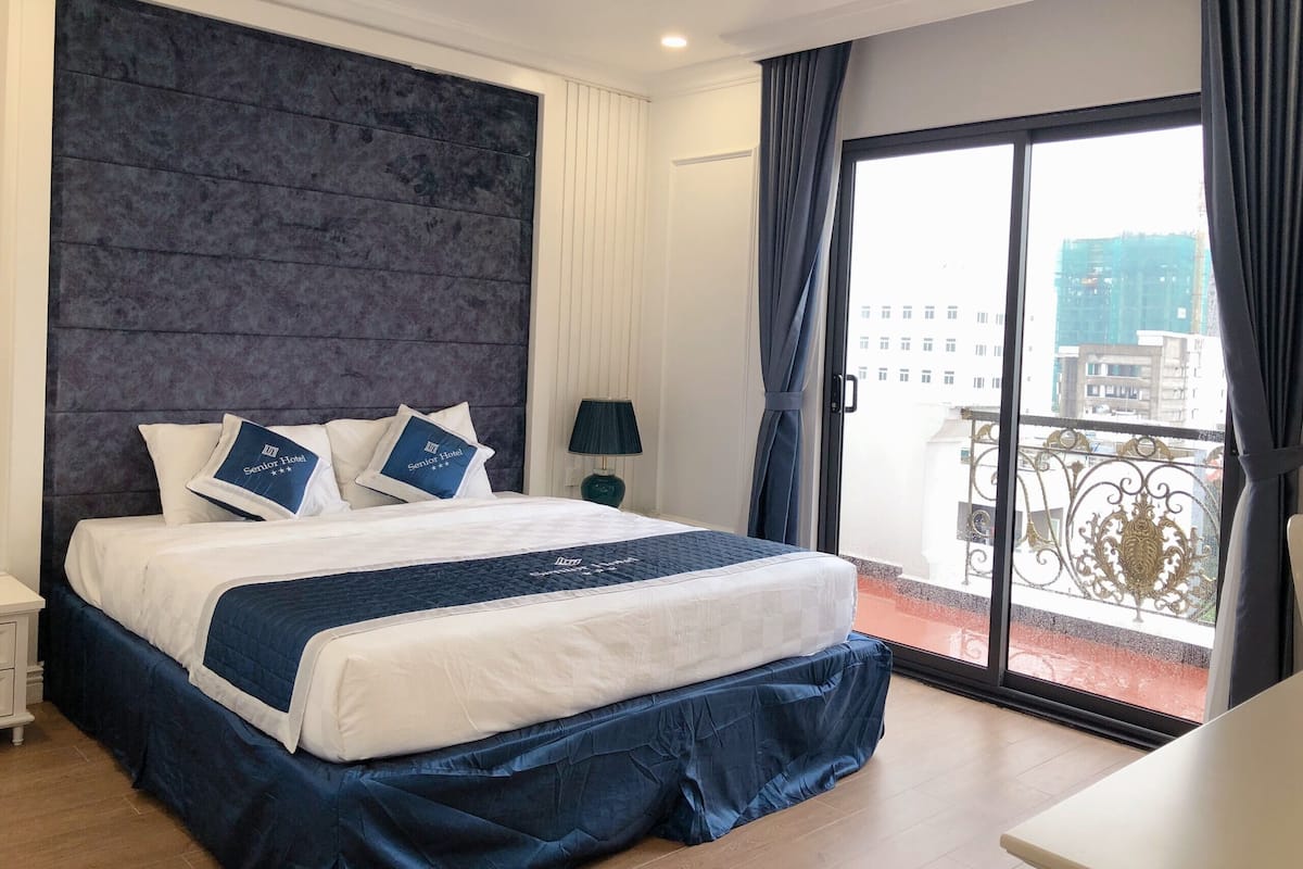 Habitación de hotel con una cama grande, una mesita de noche, una lámpara y vistas a la ciudad desde un balcón.