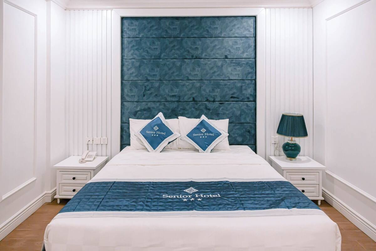Una habitación de hotel con una cama grande, dos almohadas con el logotipo del hotel, una lámpara de noche y un teléfono en la mesita.