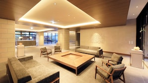 A modern hotel lobby with a central coffee table, sofas, and chairs arranged around it. There is a reception desk and a decorative wall piece. - BLISSTIA Hakone Sengokuhara (Hakone)