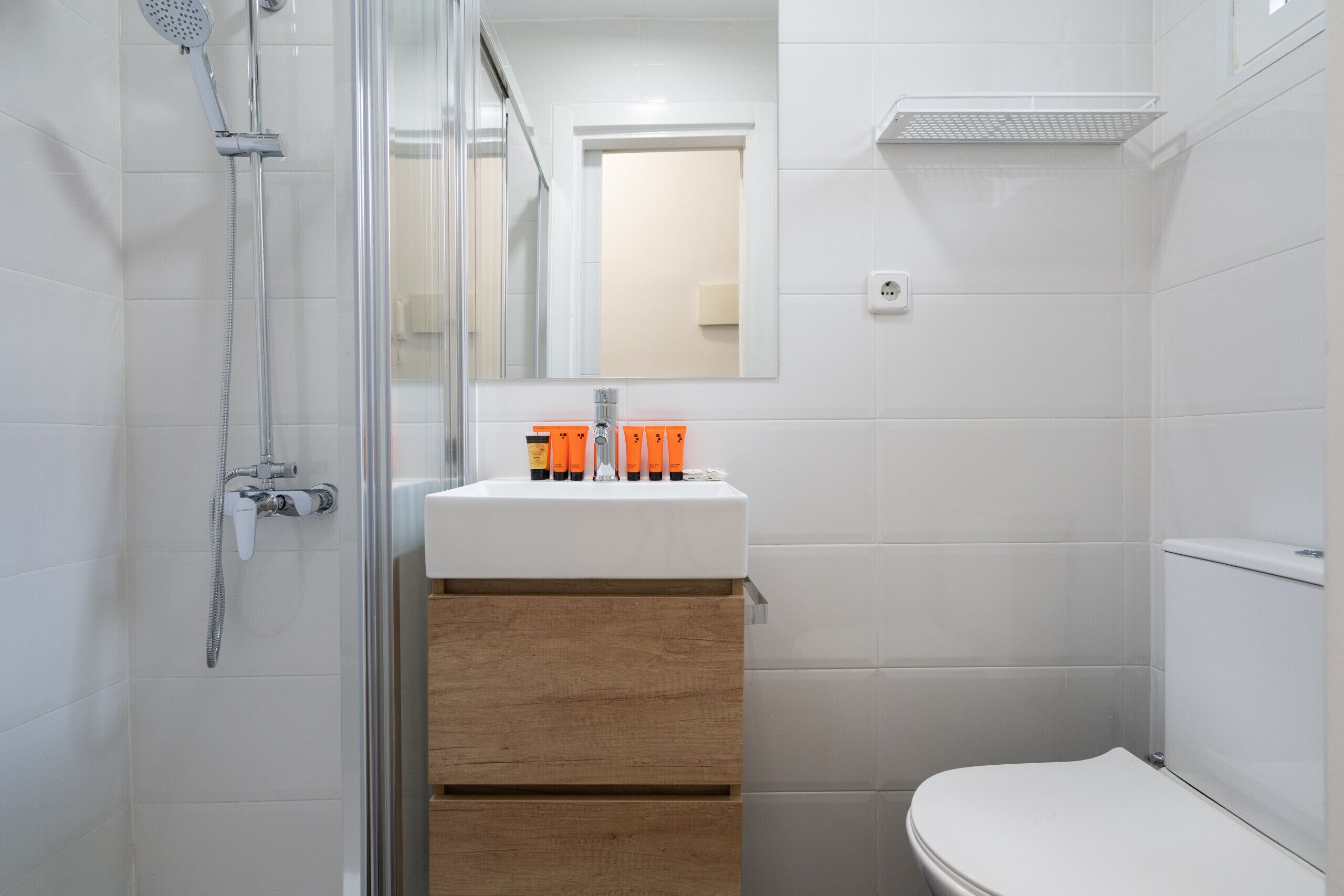 A modern bathroom with a glass shower enclosure, a white sink with orange soap dispensers, and a wooden vanity.