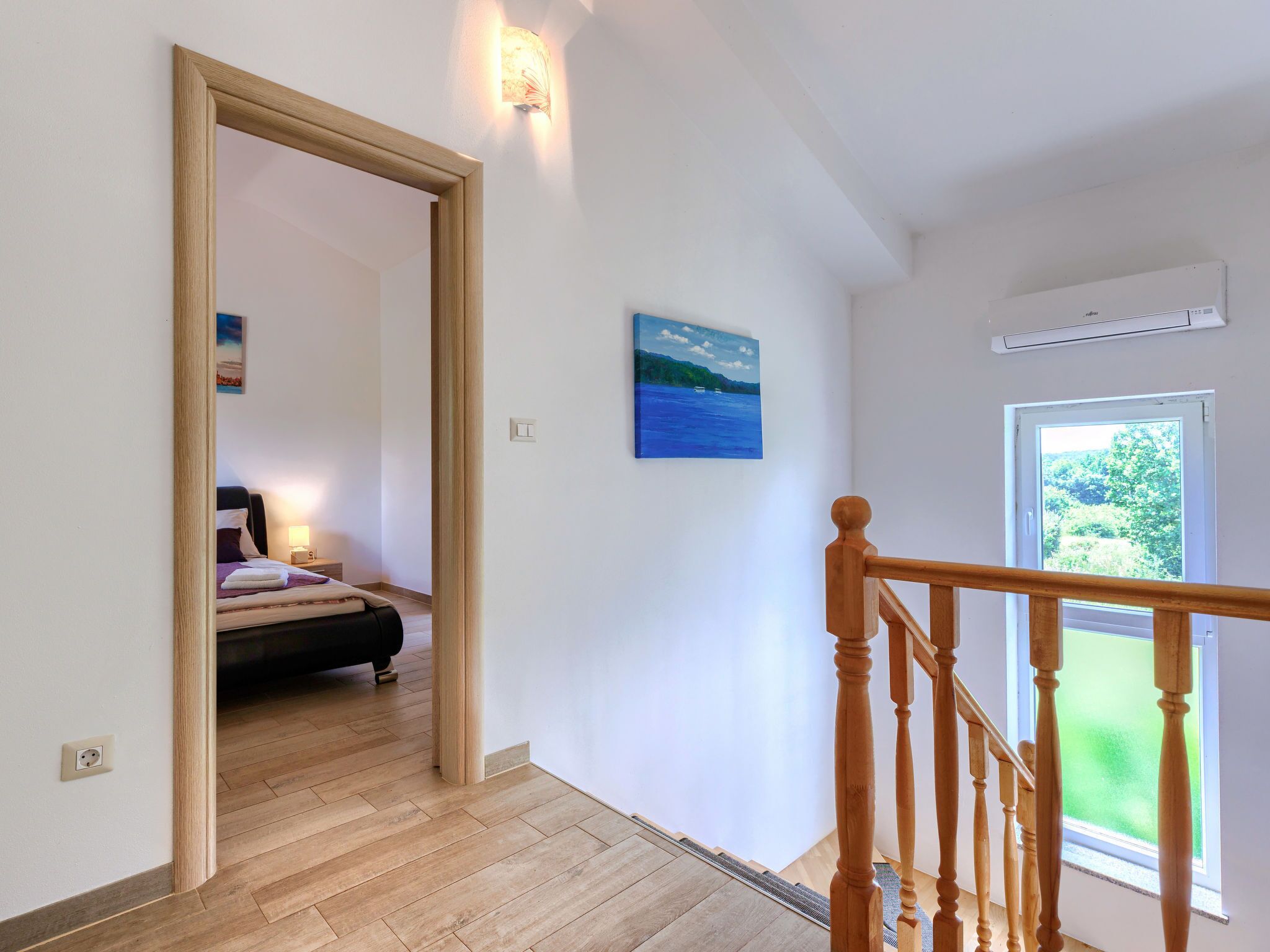 A hallway with a wooden floor, a bed in the adjacent room, a painting on the wall, and a window with a view of greenery.