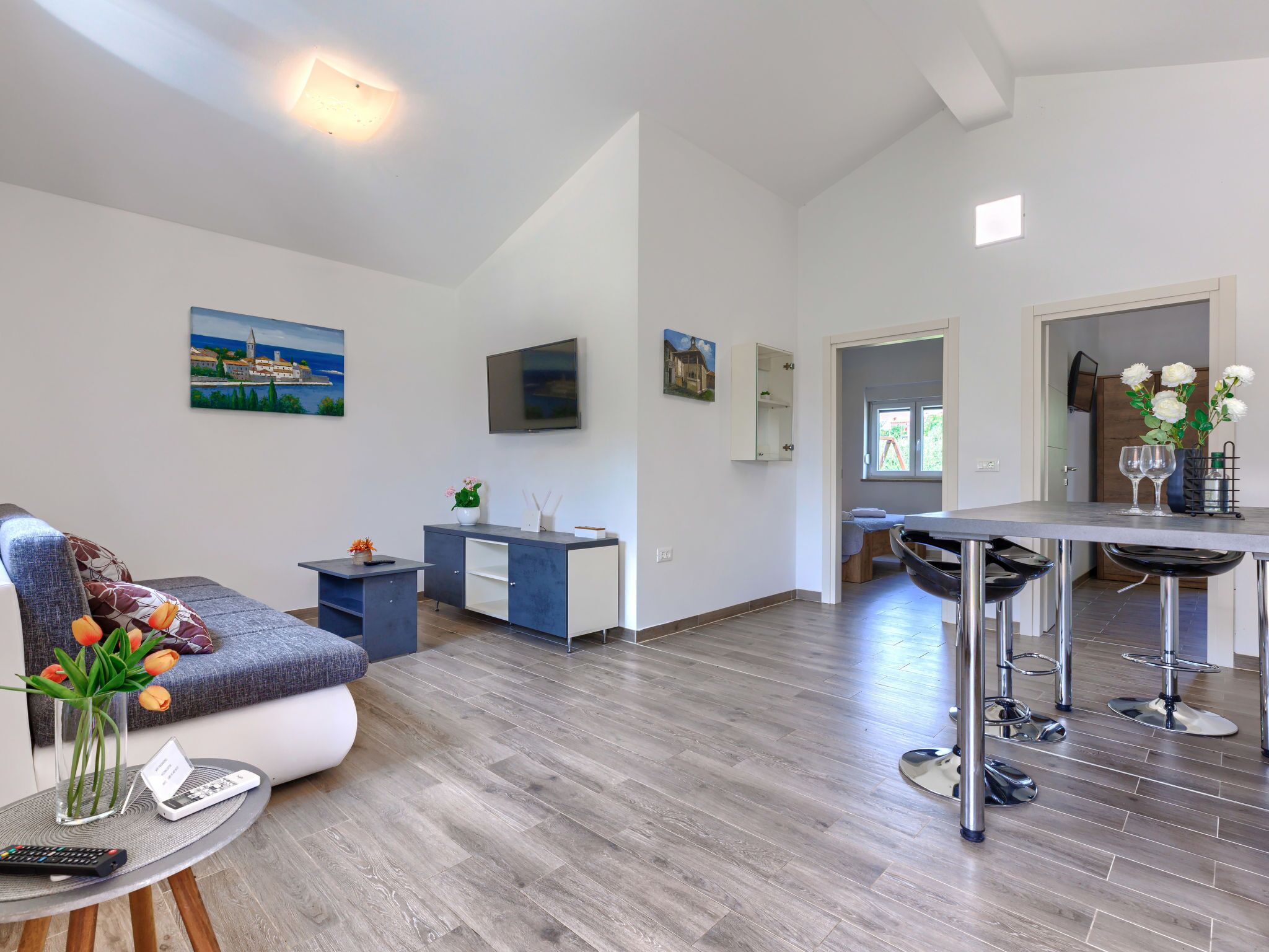 A modern living room with a sofa, a coffee table, and a TV. There is a dining area with a table and chairs. The room has wooden flooring and white walls.