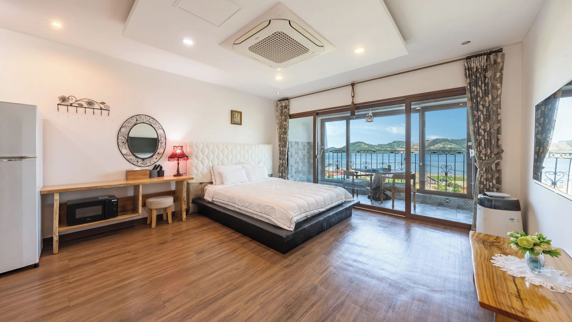 A bedroom with a bed, a desk, a chair, a mirror, and a view of the sea.