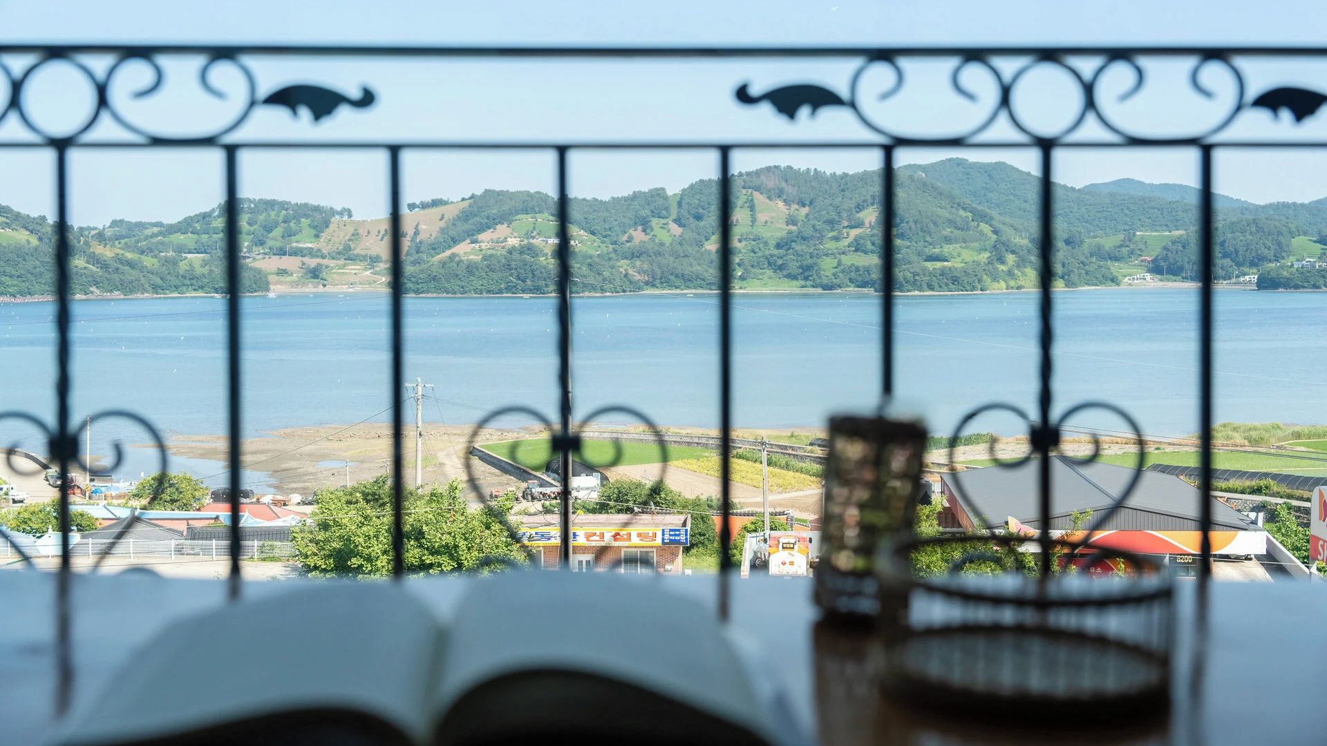 Une vue à travers une clôture en métal, avec un livre et des lunettes posés sur une table, donnant sur une étendue d’eau et un paysage vallonné.