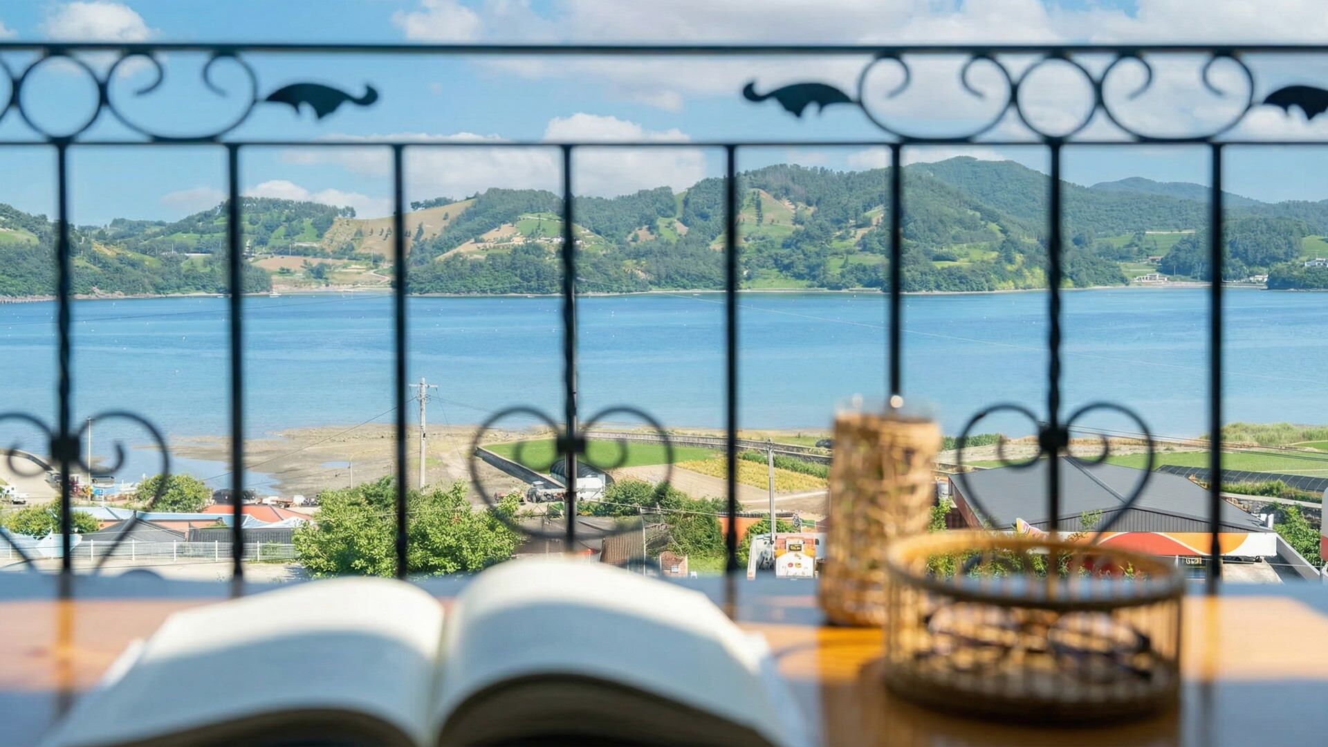 A view through a metal railing with an open book and a decorative item on a table, overlooking a body of water and green hills.
