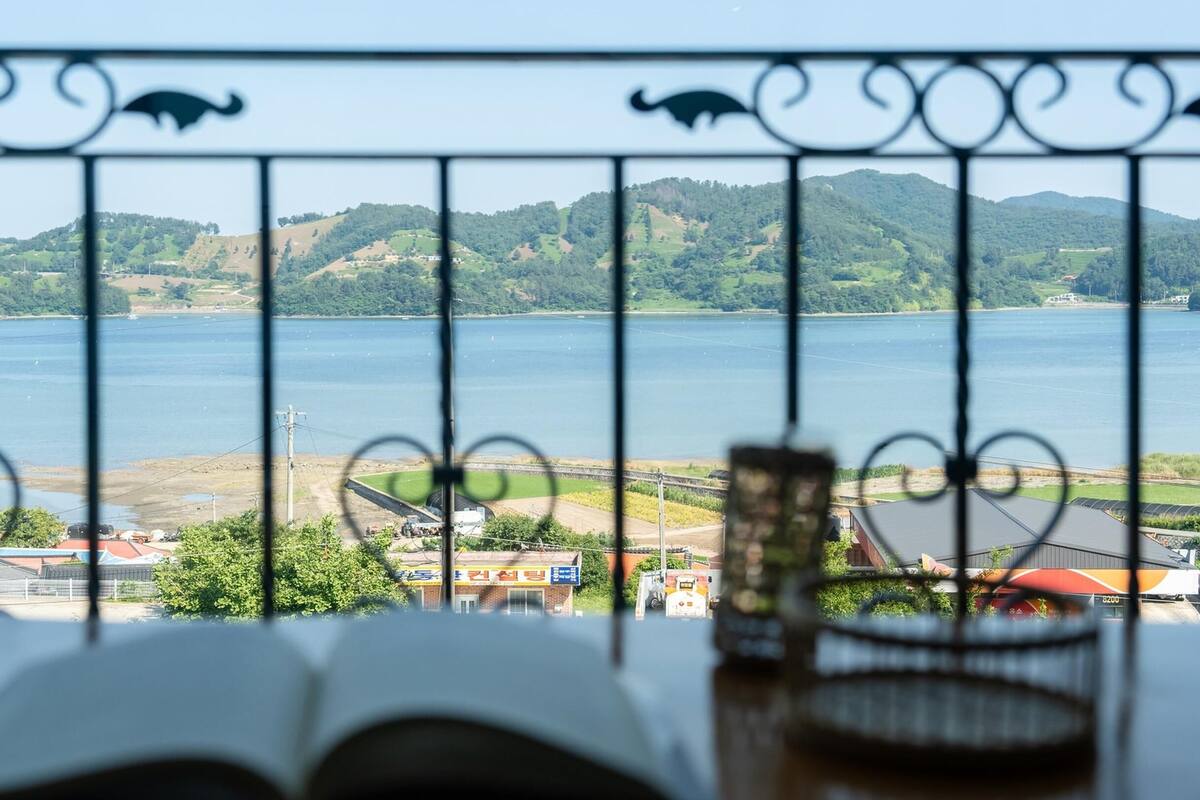 Vista a través de una cerca metálica que muestra un libro y gafas sobre una mesa, con vistas a una masa de agua y un paisaje montañoso.