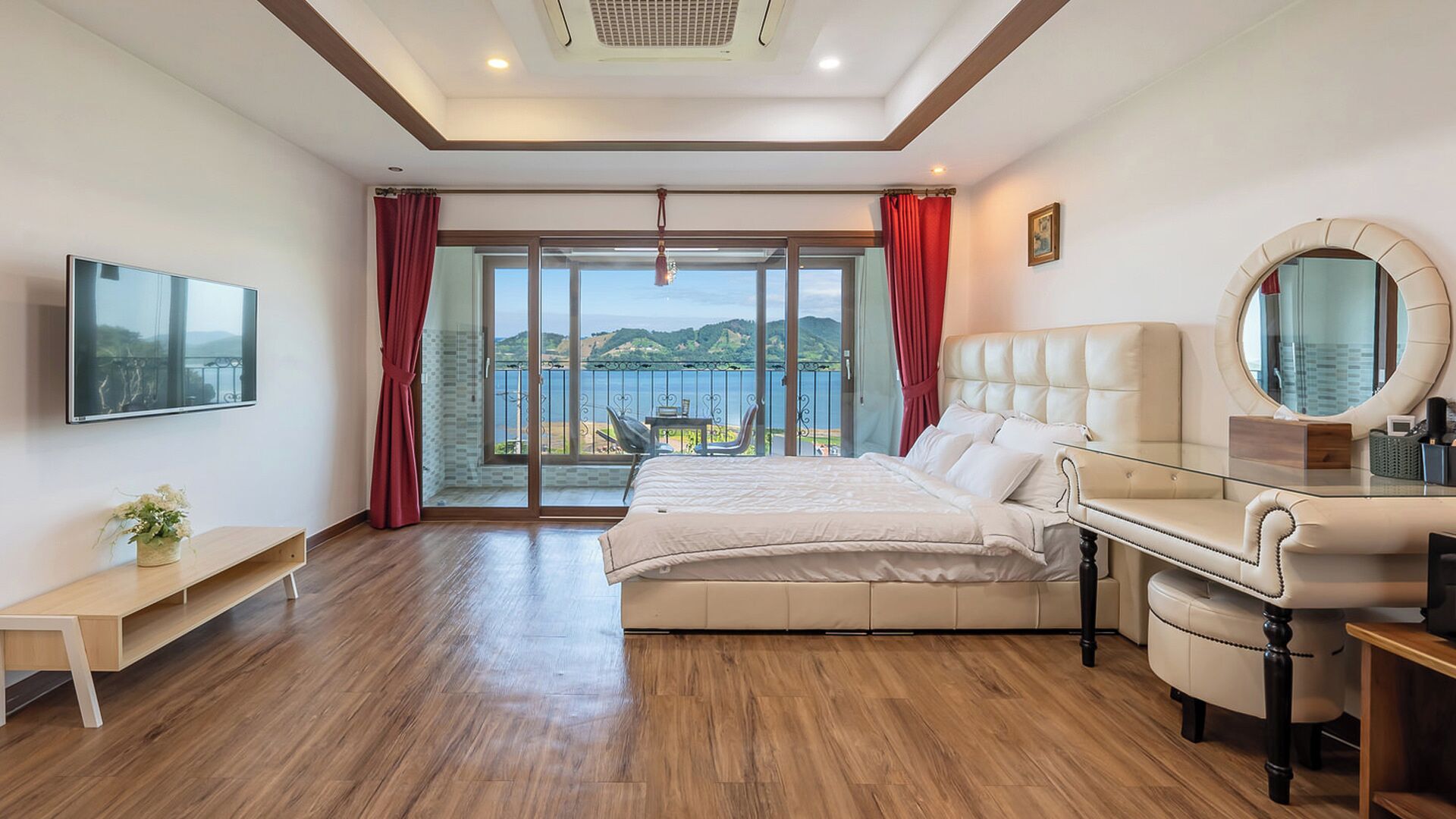 A bedroom with a large bed, a glass desk, a TV mounted on the wall, and a view of a body of water through the window.