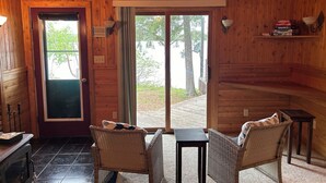 Interior acogedor de una cabaña con paredes de madera, un comedor con sillas de mimbre y una pequeña mesa, y un ventanal corredizo que da a una terraza.