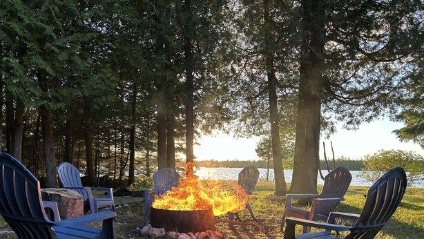 Una fogata con dos sillas Adirondack orientadas hacia ella, rodeada de árboles y un lago.