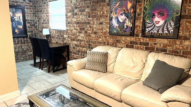 A living room with a brick wall, a beige sofa, a glass coffee table, and a dining area with a table and chairs. - Cozy 1-bedroom apartment with WiFi, AC in wonderful Baton Rouge (Baton Rouge)