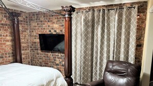 A bedroom with a brick wall, a four-poster bed, a leather armchair, and a TV mounted on the wall. - Cozy 1-bedroom apartment with WiFi, AC in wonderful Baton Rouge (Baton Rouge)