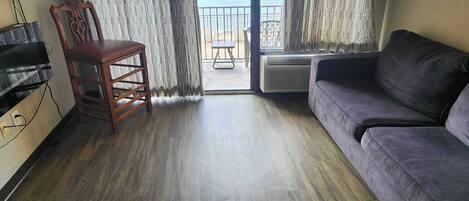 A room with a wooden floor, a grey sofa, a wooden chair, a TV, and a balcony with patterned curtains.