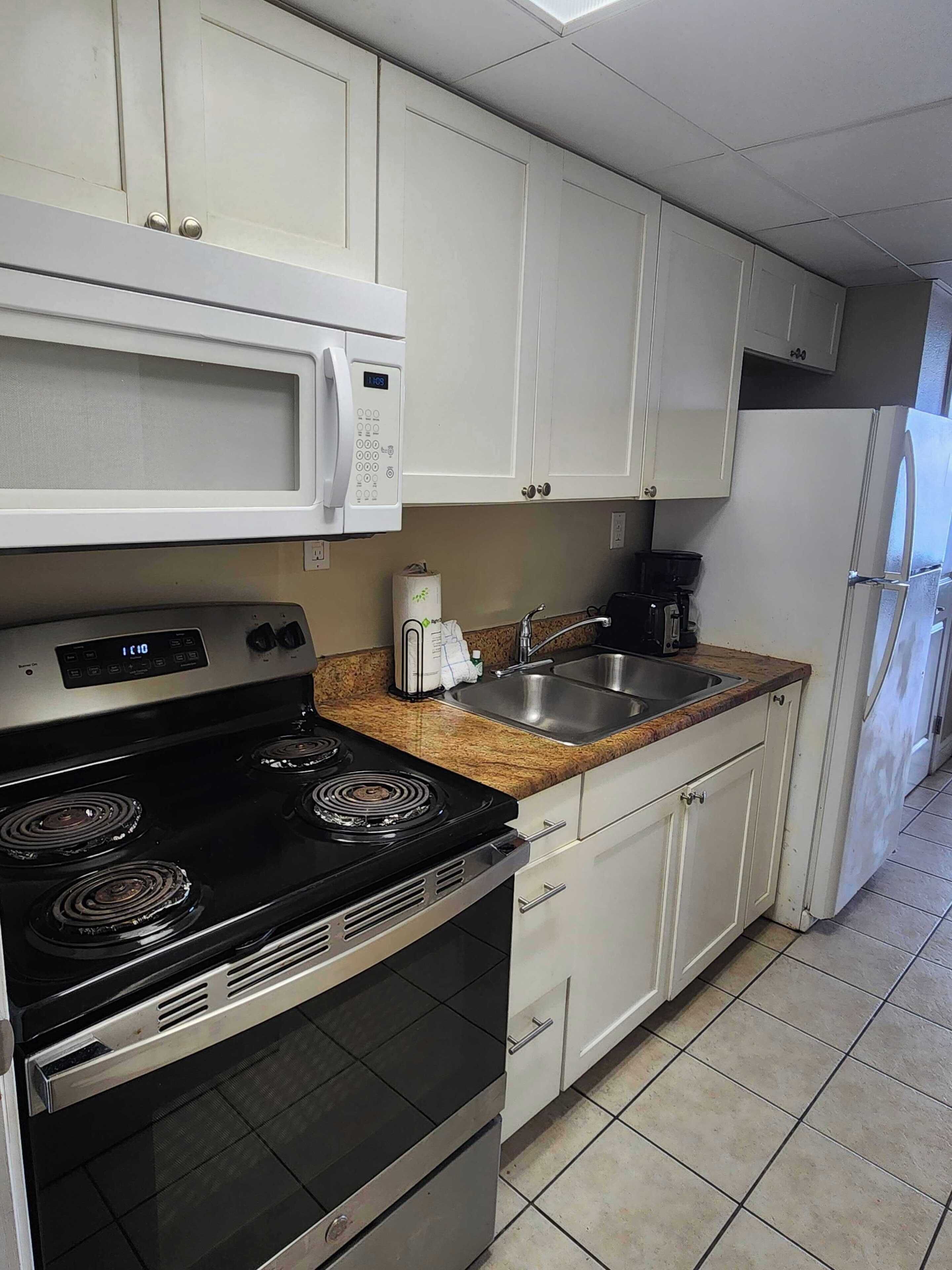 Une cuisine avec des meubles blancs, une cuisinière noire, un four à micro-ondes, un évier et un réfrigérateur.
