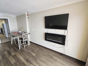 Ein modernes Wohnzimmer mit einem an der Wand befestigten Flachbildfernseher, einem Esstisch mit Stühlen und einem durch einen offenen Durchgang einsehbarer Küchenbereich.