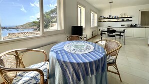 A dining area with a table and chairs, a kitchen in the background, and a view of the sea and hills. - Sa Tuna - House with Views and 30m from the Sea (Begur)