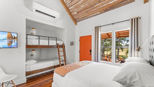 Um quarto com uma cama beliche, uma cama de solteiro, uma escada de madeira, uma janela com cortinas e um ar-condicionado fixado na parede.