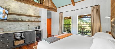 A bedroom with a large bed, a view of the outdoors through a window, a kitchenette with a microwave, and a wooden ceiling.