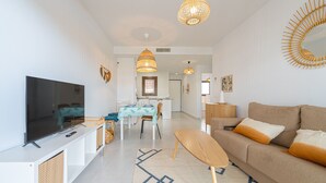 A modern living room with a flat-screen TV, a sofa, a dining table, and a kitchen in the background.