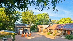 Un espace extérieur aménagé dans un style rustique, avec des tables et des chaises en bois, entouré d’arbres et bordé d’un chemin de gravier.