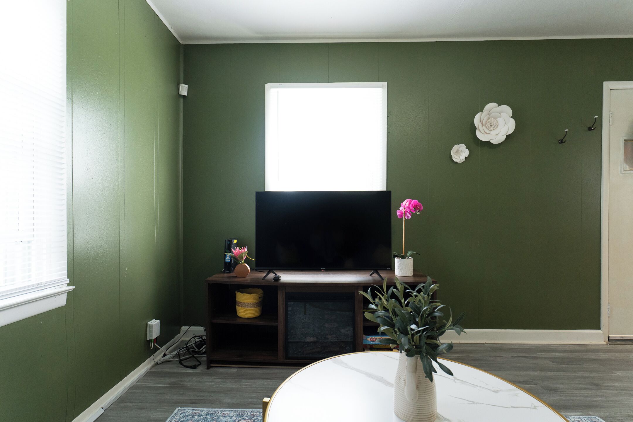 Een woonkamer met een flatscreen televisie op een houten standaard, een witte vaas met een roze bloem en een groot raam met zonwering.