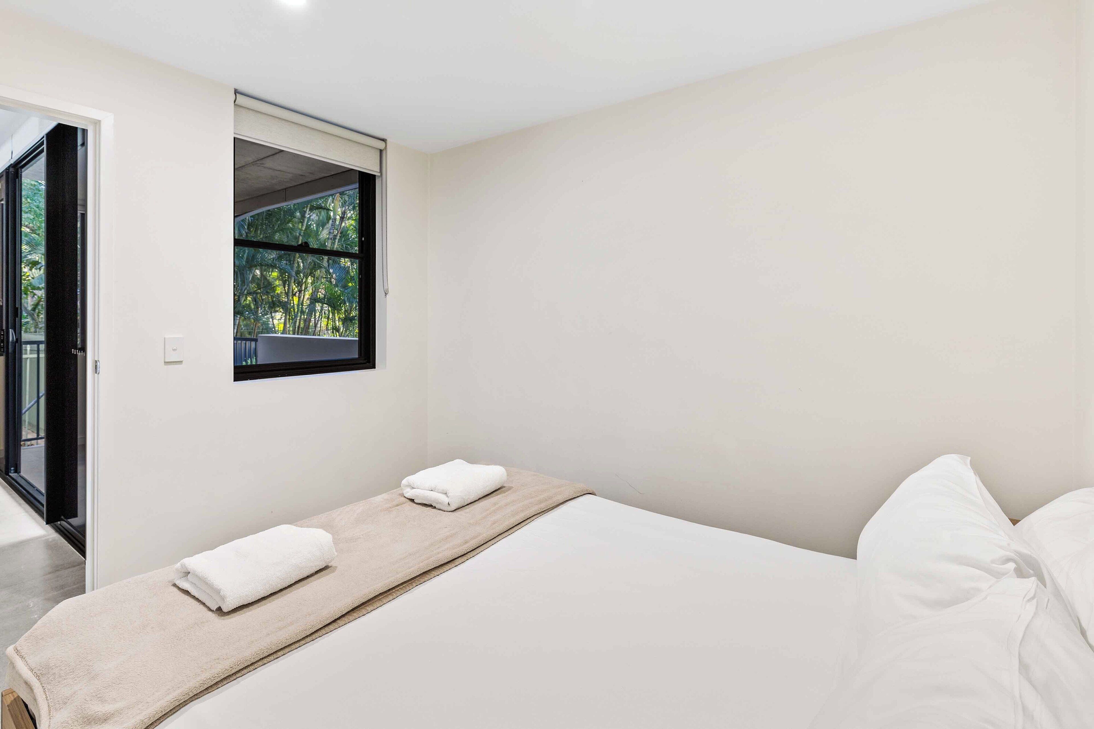 A minimalist bedroom with a single bed, a window with a view of greenery, and a door leading to an outdoor area.